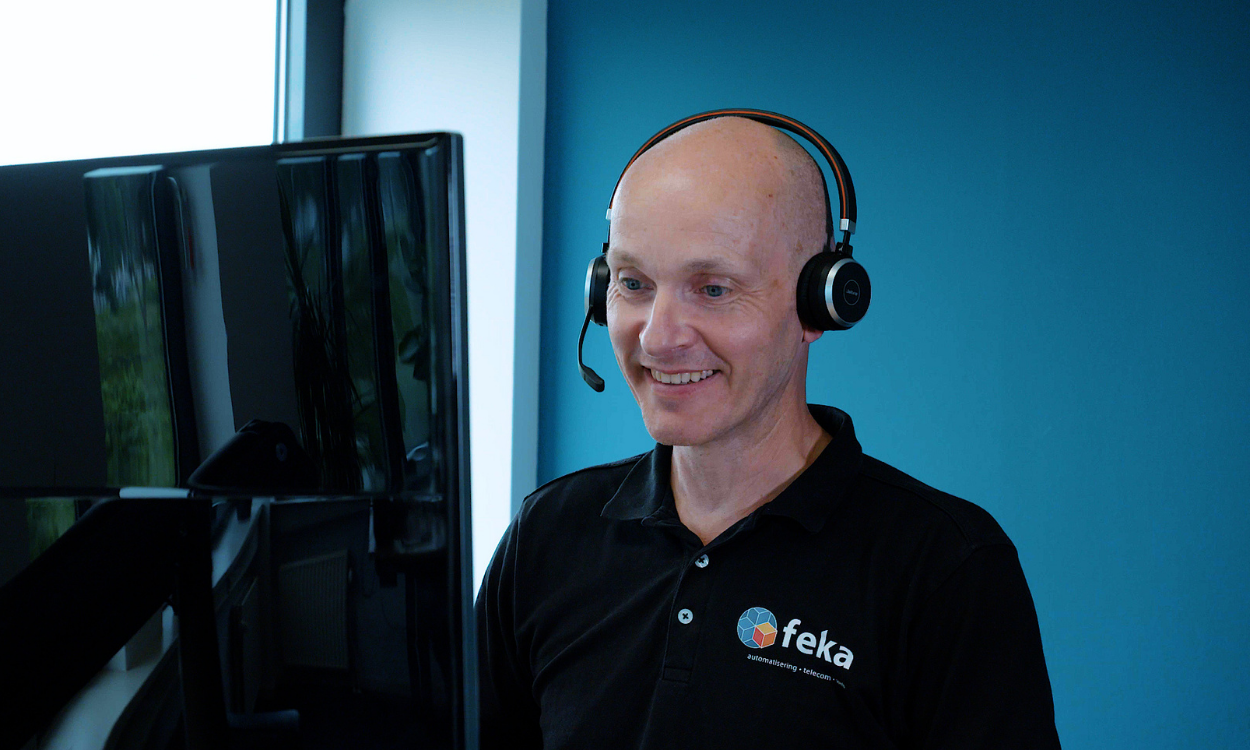 Man met headset werkt achter een computerscherm en kijkt glimlachend naar zijn scherm in een moderne kantoorruimte.